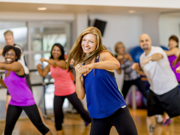 A woman is leading a multi-ethnic boxing class, everyone is wearing athletic clothing and is throwing a punch together - they are smiling and looking at the camera.