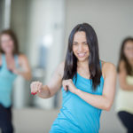 A multi-ethnic group of young adult women are at the gym taking a dance fitness class together. They are moving to the music and are smiling while looking at the camera.