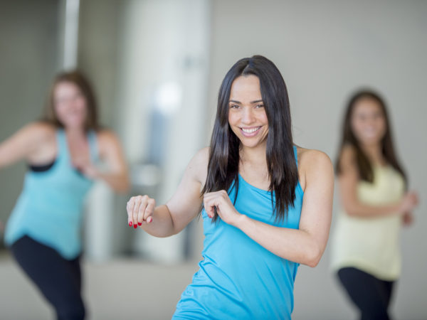 A multi-ethnic group of young adult women are at the gym taking a dance fitness class together. They are moving to the music and are smiling while looking at the camera.