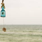 An empty, aqua-blue bottle hanging with a seashell as a summer wind chime decoration and wind chime - shot in an afternoon with sea coast in the background.
