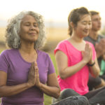 A senior woman of Pacific Islander descent meditates during a group yoga class. She has a slight smile and is seated in the prayer pose. The multi-ethnic group is outdoors. Men and woman of different ages are taking the class. The class participants are seated in a line and they are all doing the prayer pose.