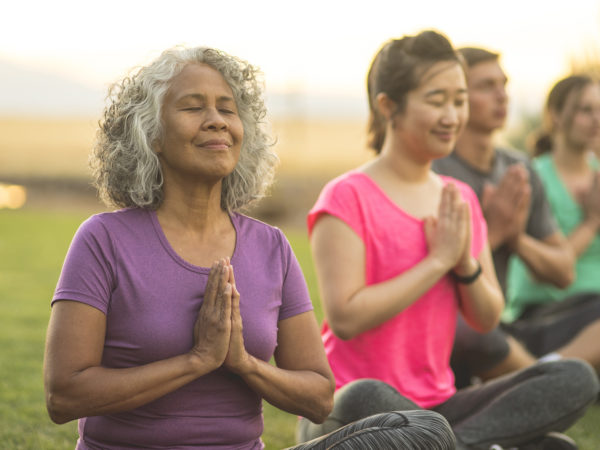 A senior woman of Pacific Islander descent meditates during a group yoga class. She has a slight smile and is seated in the prayer pose. The multi-ethnic group is outdoors. Men and woman of different ages are taking the class. The class participants are seated in a line and they are all doing the prayer pose.