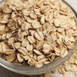 Close-up of a small glass bowl filled with rolled oats.  The oats are overflowing out of the bowl and have spilled onto the surface next to the bowl.  The bowl has rings around the edge, and the oats are raw and in a dry form.