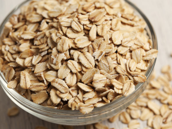 Close-up of a small glass bowl filled with rolled oats. The oats are overflowing out of the bowl and have spilled onto the surface next to the bowl. The bowl has rings around the edge, and the oats are raw and in a dry form.