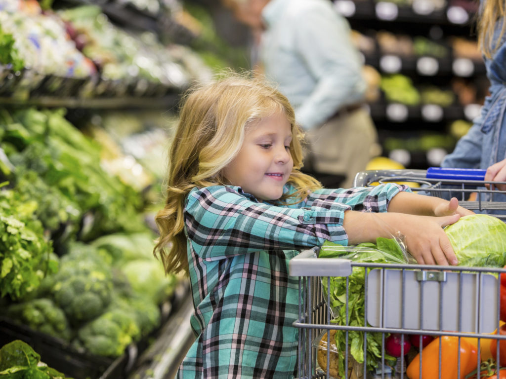 Elementary age Caucasian blonde little girl is smiling while placing lettuce in shopping cart. Child is shopping for produce and healthy food with her mother in local grocery store. Child is wearing a plaid shirt. Other customers are shopping in background.