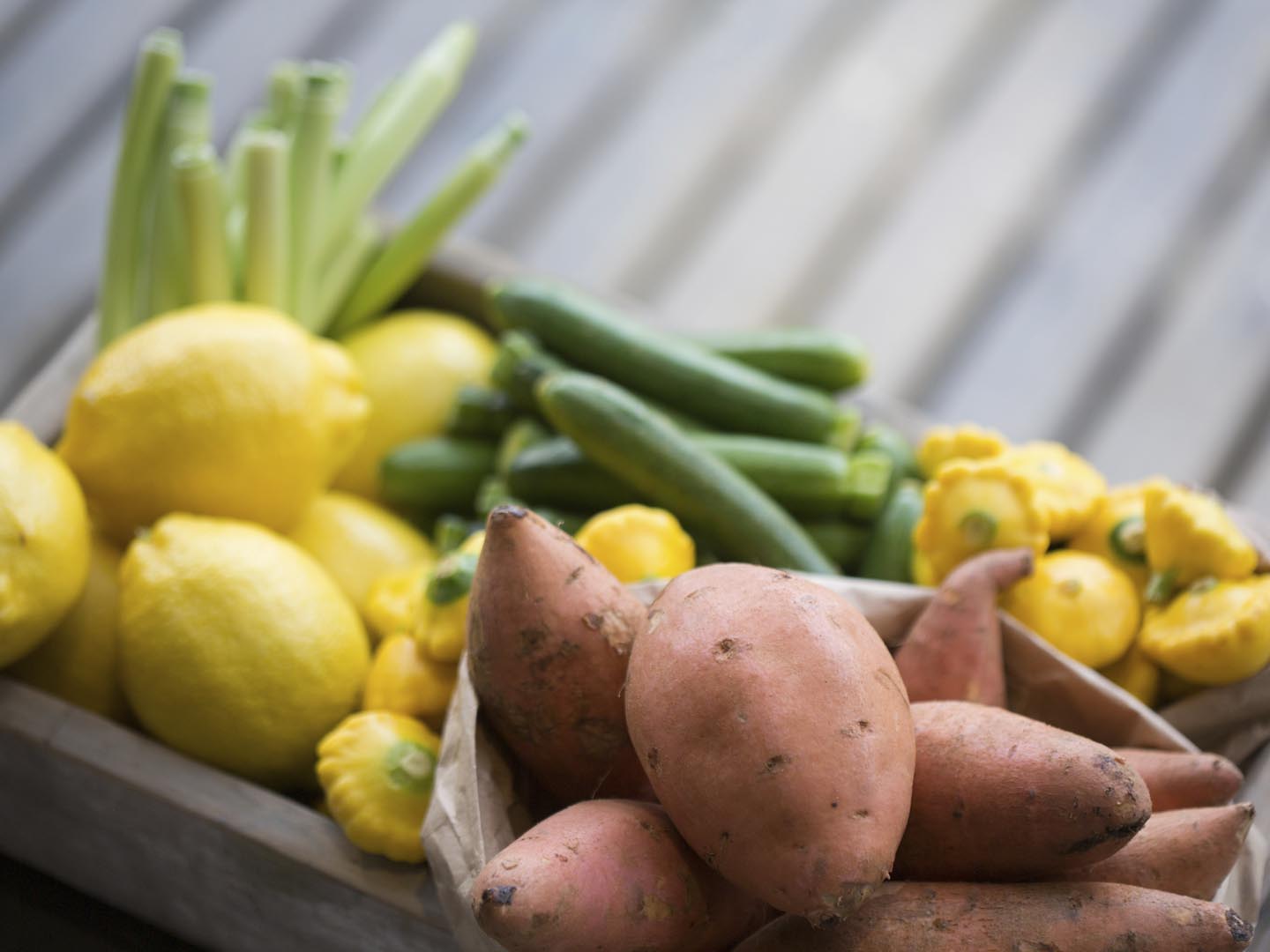 A horizontal composition of a small wooden crate filled with fruit and vegetables, including lemons, lemon grass, courgettes, pattypans and red sweet potatoes