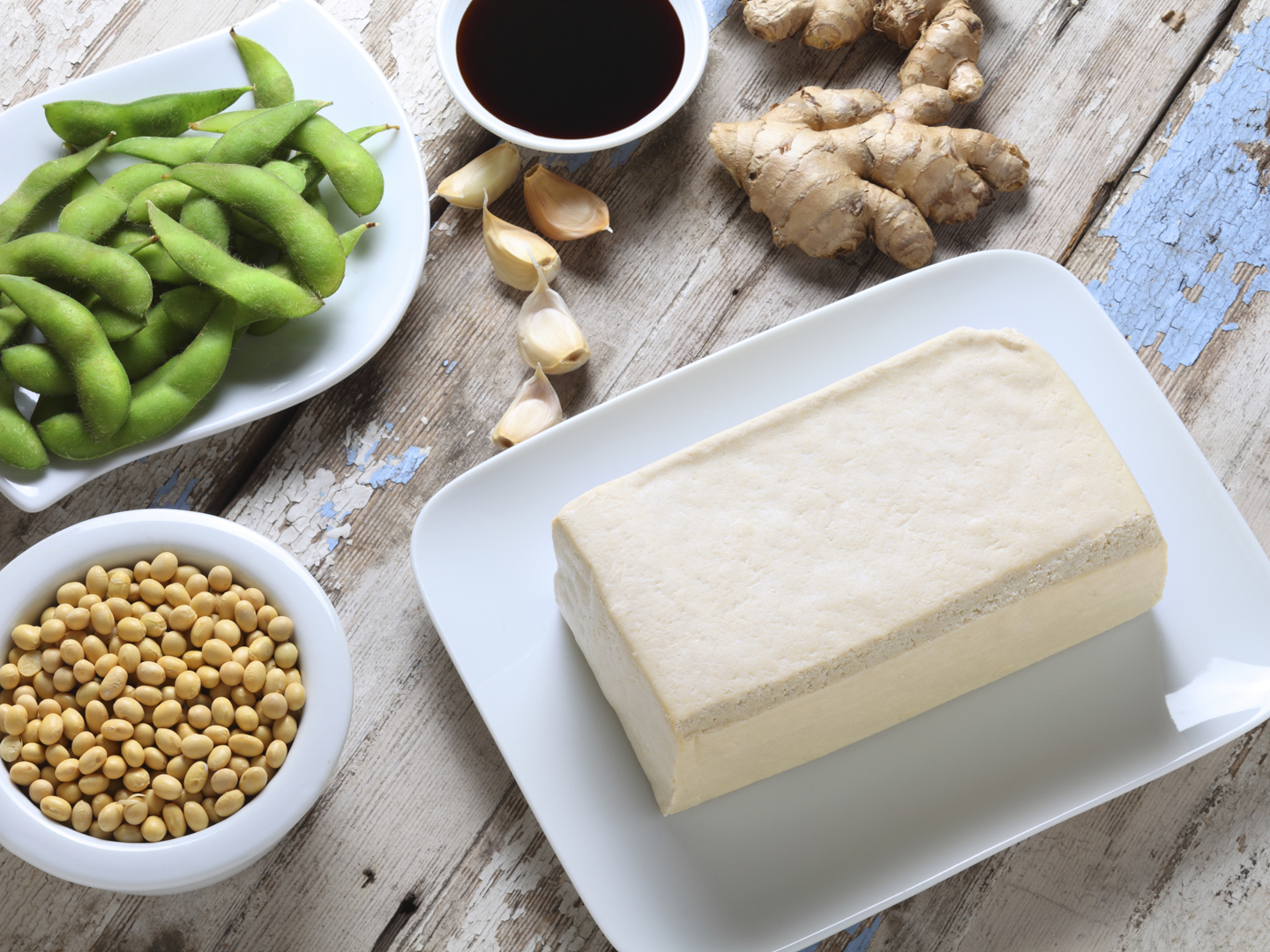 A block of tofu with a bowl of edamame, a bowl of soy beans, garlic cloves,soy sauce, and ginger root.