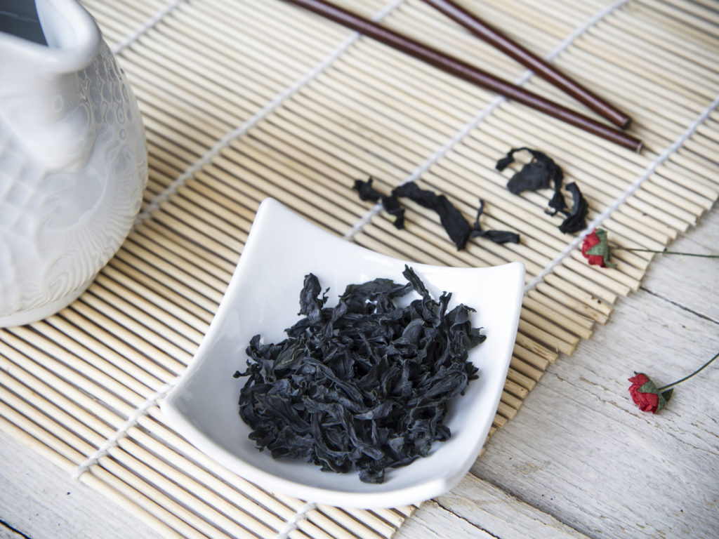 Horizontal color photo, with seaweed wakame dehydrated in a small white bowl, white jug of water to hydrate algae, chopsticks and bamboo placemat.