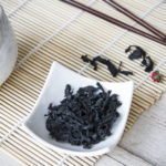 Horizontal color photo, with seaweed wakame dehydrated  in a small white bowl, white jug of water to hydrate algae, chopsticks and bamboo placemat.