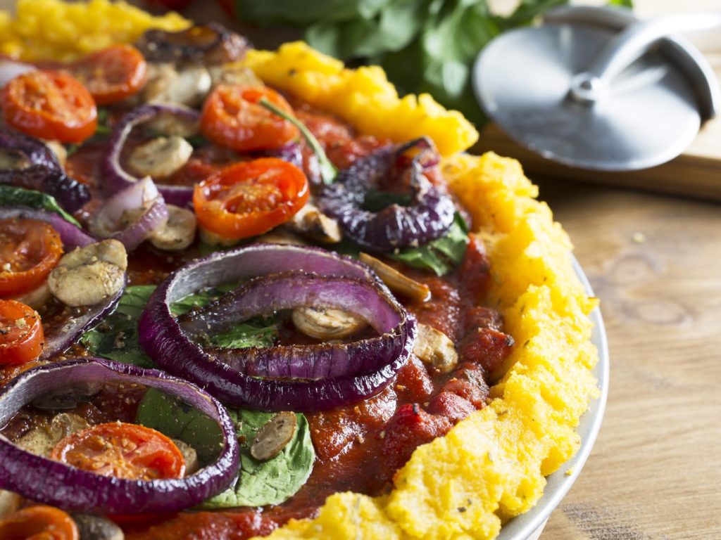 Whole vegan polenta pizza with cherry tomatoes, mushrooms, spinach and red onions. Close up with pizza slicer out of focus in the background, horizontal orientation.
