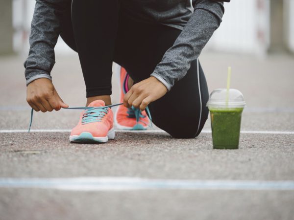 Fitness workout and healthy nutrition concept.  Detox smoothie drink and running footwear close up. Female athlete tying sport shoes laces before training outdoor.