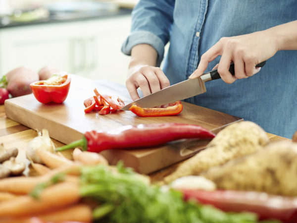 Midsection image of woman chopping red bell pepper on cutting board. There are various vegetables on wooden table. Focus is on her hands. Female is preparing food. She is in domestic kitchen.
