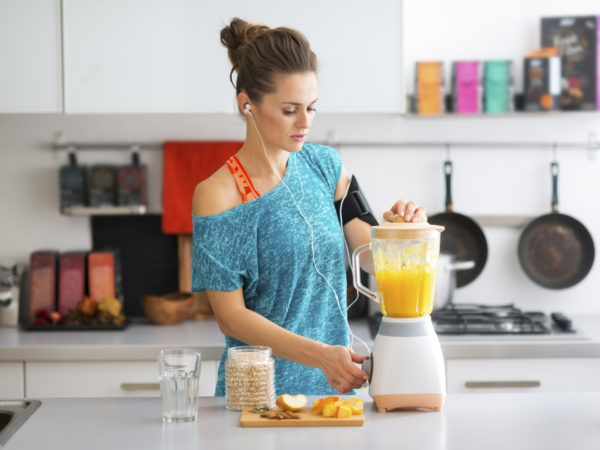 A sporty woman is standing in her kitchen, making a smoothie with fresh, seasonal fruits, nuts, and oats, to complete her healthy start to the morning.