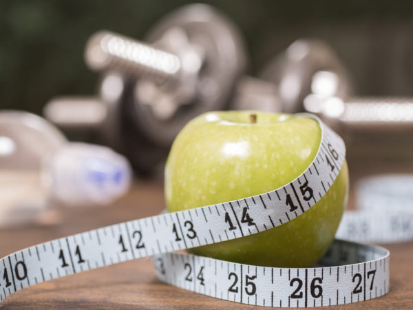 Fitness, exercise, dieting themed scene with green apple and tape measure in foreground with barbells and water bottle in background.  Group of objects lie on wooden surface in gym or health club setting with outdoor view in far distance.