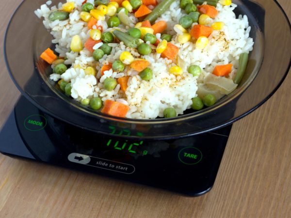 Translucent white plate with rice and vegetables is at home kitchen electronics scales to count calories in food on wooden table. Photo closeup