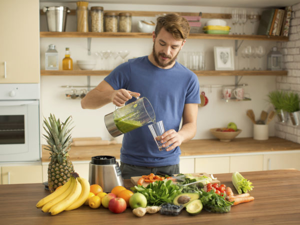Young man preparing fruit or vegetable smoothie or juice in his kitchen. Different fruits and vegetables on the table. Pouring green smoothie in glass. Caucasian, casual style, brown hair and beard.