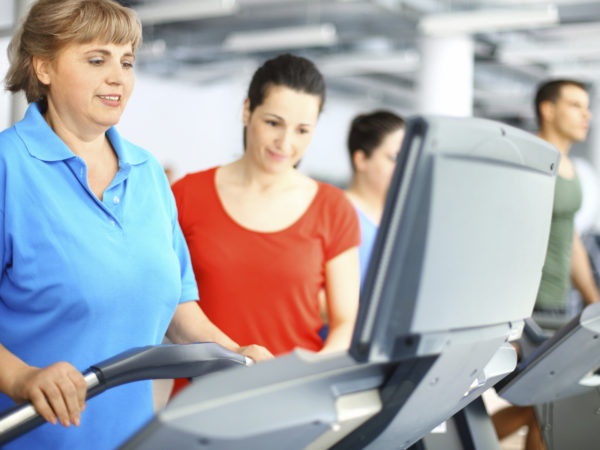Overweight senior  caucasian woman doing cardio workout on a treadmill in local gym with her coach assisting and supporting.She&#039;s wearing blue t-shirt and has her mid long partially gray hair.Coach is standing next to her and wearing red tank top.There&#039;s blurry muscular guy in background