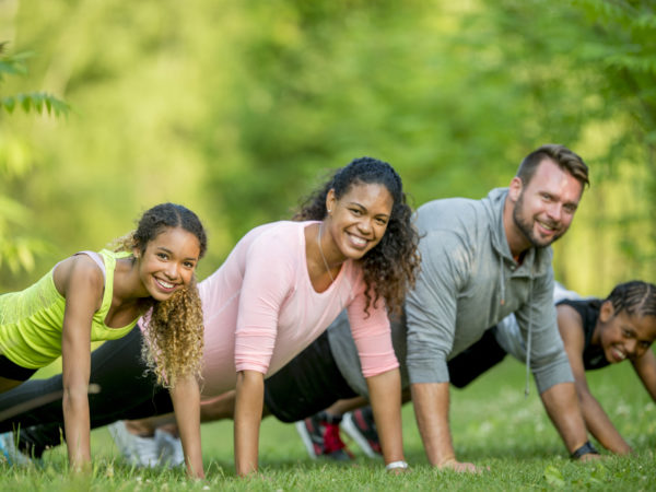 A multi-ethnic family is exercising together outside at the park, they are holding the plank position and are smiling while looking at the camera.