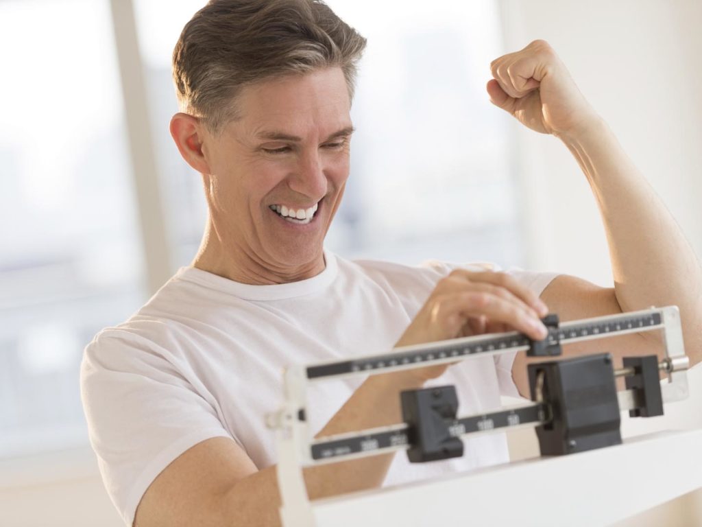 Excited mature man clenching fist while using balance weight scale at gym
