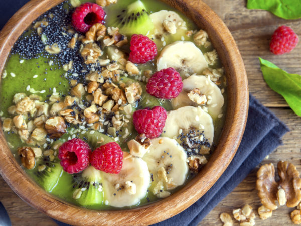 Healthy breakfast green smoothie bowl topped with fruits, nuts, berries and seeds over wooden background close up