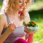 Young slim woman holding a bowl of fresh salad ofter working out outdoors in the park. Eating on fresh air, sitting on the grass and enjoying early morning. Healthy lifestyle.