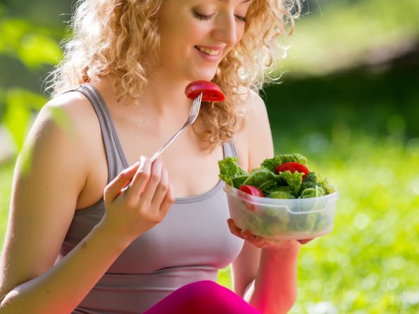 Young slim woman holding a bowl of fresh salad ofter working out outdoors in the park. Eating on fresh air, sitting on the grass and enjoying early morning. Healthy lifestyle.