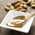 A dish of homemade almond butter on rustic wood surface.  Whole almonds in background and wooden spoon with fresh almond butter in foreground.