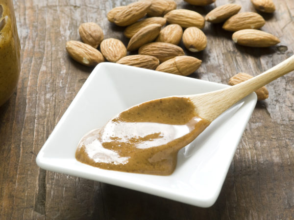 A dish of homemade almond butter on rustic wood surface.  Whole almonds in background and wooden spoon with fresh almond butter in foreground.