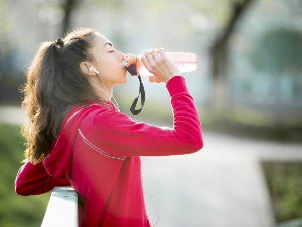 Profile portrait of sporty woman drinking in park after jogging. Female athlete runner getting ready for running routine. Fit girl listening music and enjoying drink with closed eyes outdoors
