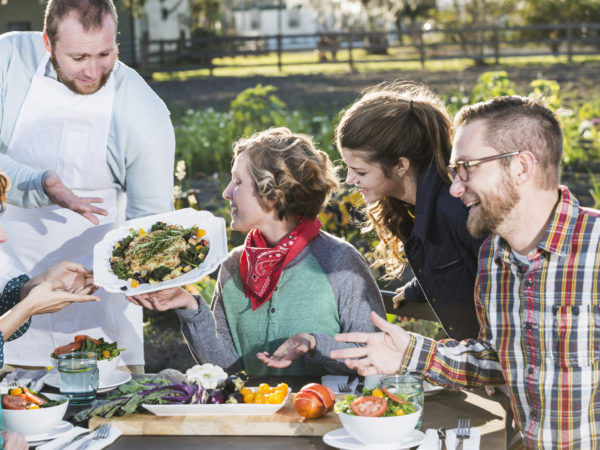 A group of people sitting outdoors at a dining table, being served locally grown food.  The chef or waiter is standing, presenting a dish of beautifully prepared vegetables.  This image represents the farm-to- table-or-farm to fork movement.