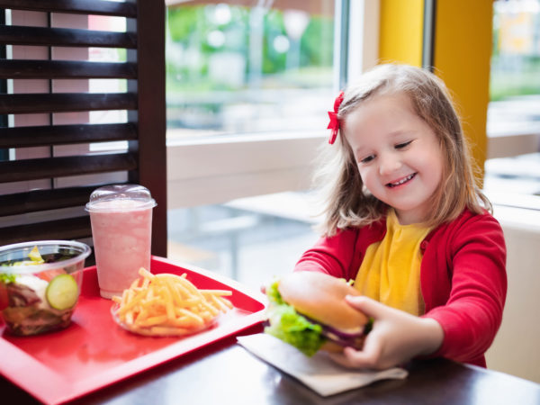 Little girl eating hamburger and French fries in a fast food restaurant. Child having sandwich and potato chips for lunch. Kids eat unhealthy fat food. Grilled fastfood sandwich for children.