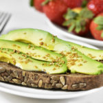 closeup of an avocado toast made with rye bread and a bowl full of strawberries in the background