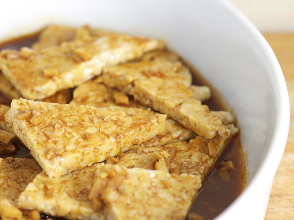 "Pieces of tempeh marinating in garlic, ginger, oil and spices in a white baking dish"