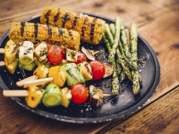 High angle view of a healthy selection of grilled vegetable kebabs, asparagusand corn on a dark plate on a wooden surface