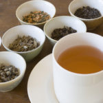 Selection of dried tea leaves from around the world and a cup of hot brewed tea, ready for comparative tasting and sipping. The white saucer holds a full pour of the beverage. Chinese, Japanese, and Indian varieties such as oolong, jasmine, darjeeling, and other black and green choices are shown.