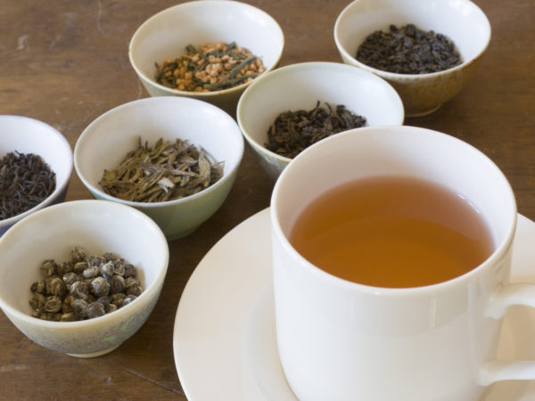 Selection of dried tea leaves from around the world and a cup of hot brewed tea, ready for comparative tasting and sipping. The white saucer holds a full pour of the beverage. Chinese, Japanese, and Indian varieties such as oolong, jasmine, darjeeling, and other black and green choices are shown.