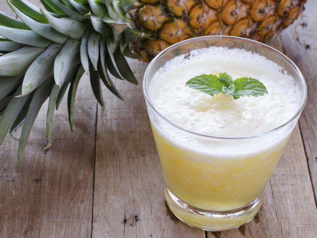 A glass of pineapple smoothie with fresh mint leaves and pineapple on wooden table.