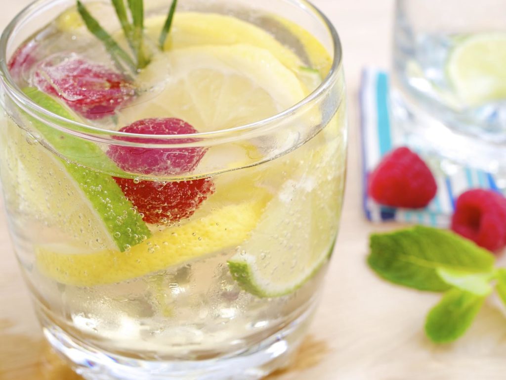 Ein glas mit eiskalter frischer Limonade, Limetten- und Zitronenscheiben und Eisw?rfeln auf einem Holztisch neben einem zweiten leeren glas, garniert mit einem st?ck frischer Minze und Himbeeren.