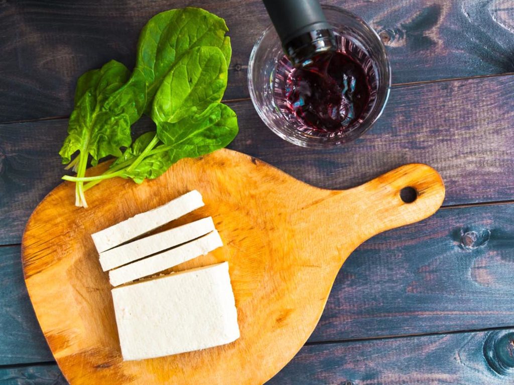 Slices of raw tofu on shabby cutting board, green leaves of fresh spinach and glass of red wine filling from the bottle