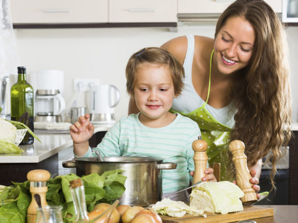 Young mother with little daughter cooking soup together at domestic kitchen. Focus on girl