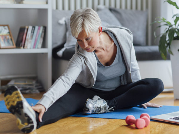 Mature woman exercising at home, learning exercises online with laptop. Sitting on floor and stretching.