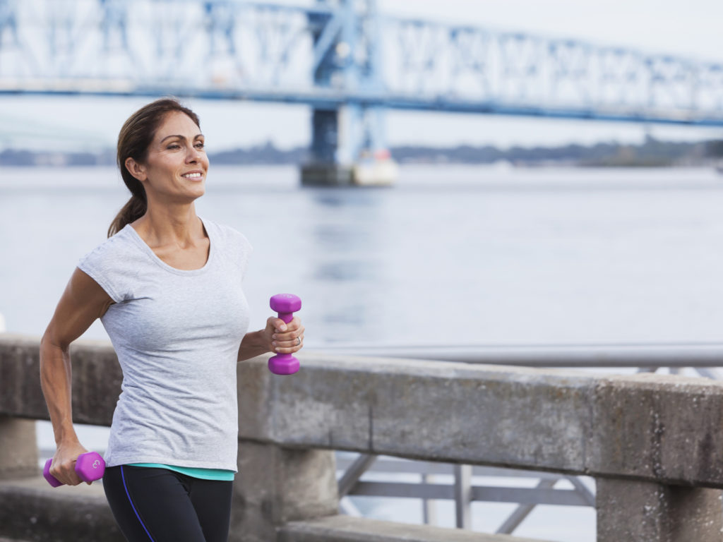Mature woman staying fit, running or power walking along the waterfront, carrying dumbbells in her hands.