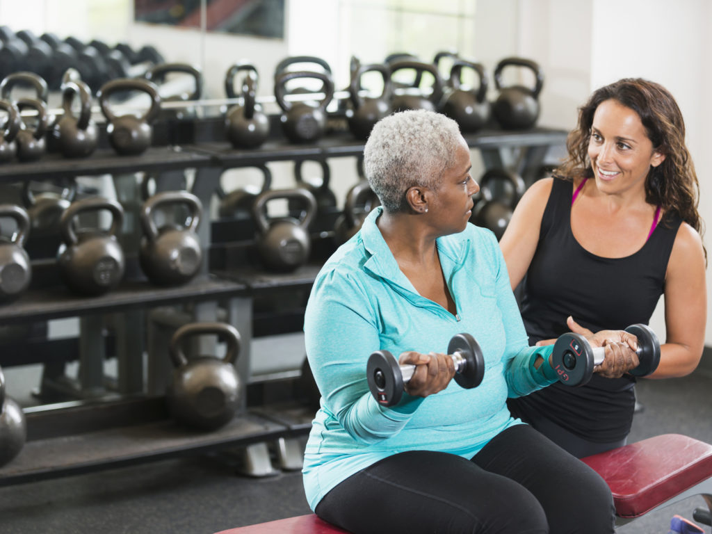 Friendly personal trainer at a fitness center helping a senior African American woman. They are talking face to face, sitting on a bench while the senior lifts dumbbells in her hands to strengthen her arms and biceps. There are kettle bells on the shelf in the background.