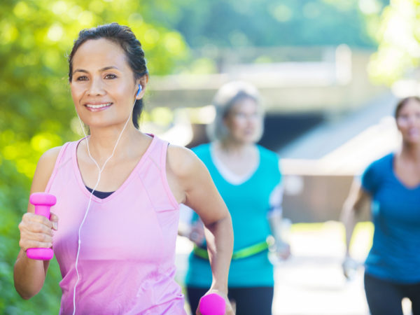 Asian senior woman uses pink handweights while power walking in the park. She is wearing a pink tank top. She has brown hair pulled back into a ponytail. She is wearing earphones and listening to musics. Women are walking behind her.