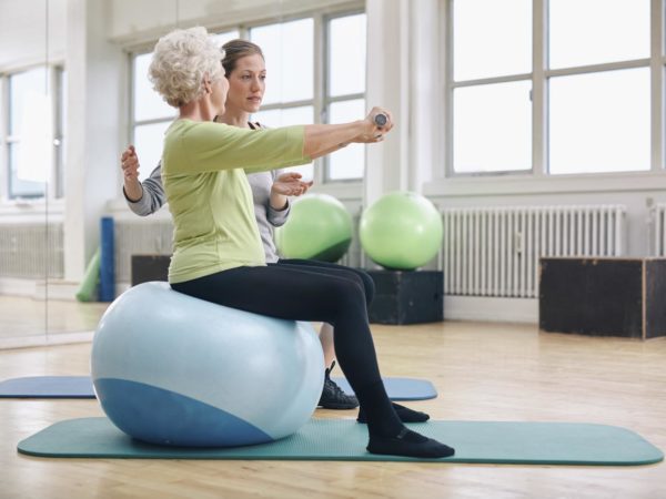 Female trainer assisting senior woman lifting weights in gym. Senior woman sitting on pilates ball doing weight exercise being assisted by personal trainer at health club.