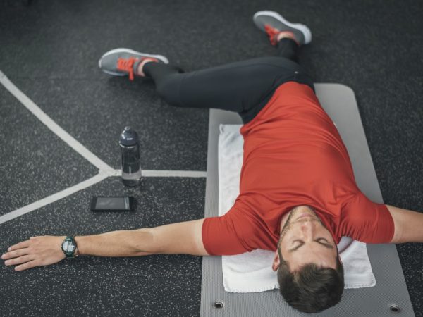 Sporty man stretching back before gym workout. Fitness strong male athlete on floor mat and towel warming up with bottle and cellphone.