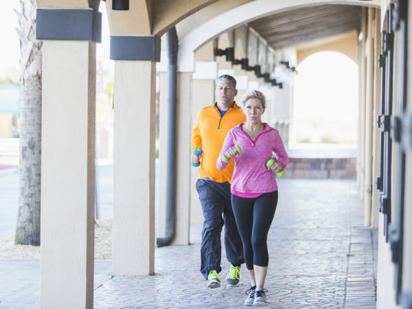 A mature couple exercising together, running or jogging with handweights, under the archways of a building. The woman is running a few steps in front of the man.