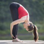 Fit young beautiful woman wearing red tank top and black sporty leggings working out outdoors in park on summer day, doing Standing Head to Knees posture, Uttanasana pose, full length