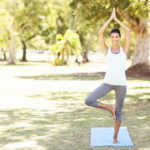 Full length portrait of smiling woman performing yoga in tree pose at park. Horizontal shot.