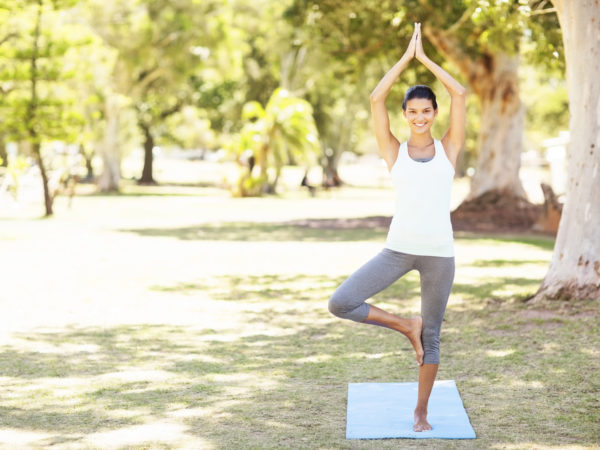 Full length portrait of smiling woman performing yoga in tree pose at park. Horizontal shot.